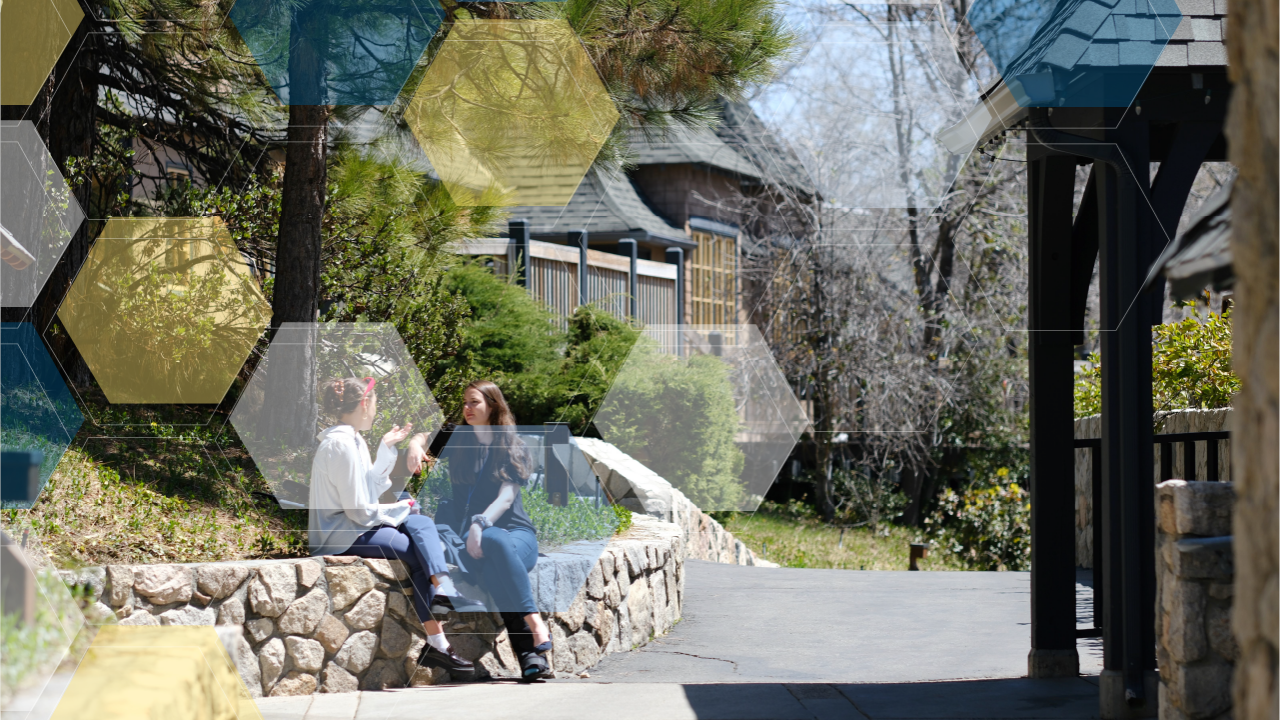 Two women sit on a stone wall surrounded by trees and wooden buildings.