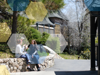 Two women sit on a stone wall surrounded by trees and wooden buildings.